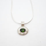 A silver pendant necklace featuring a textured, organic-shaped silver bezel holding a round green gemstone, suspended from a fine silver chain against a plain white background.