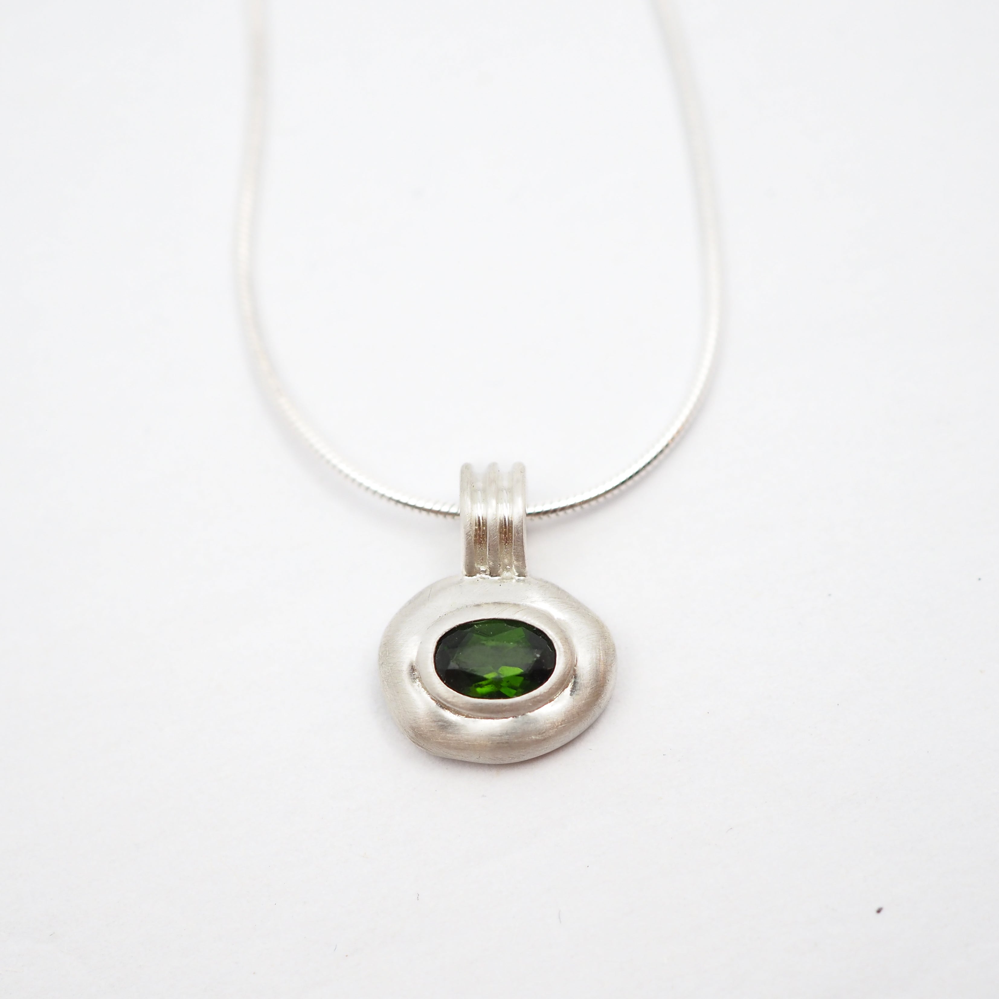 A silver pendant necklace featuring a textured, organic-shaped silver bezel holding a round green gemstone, suspended from a fine silver chain against a plain white background.