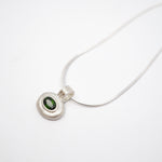 A silver pendant necklace featuring a textured, organic-shaped silver bezel holding a small oval green gemstone, suspended from a delicate silver snake chain on a plain white background.