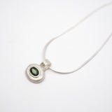 A silver pendant necklace featuring a textured, organic-shaped silver bezel holding a small oval green gemstone, suspended from a delicate silver snake chain on a plain white background.