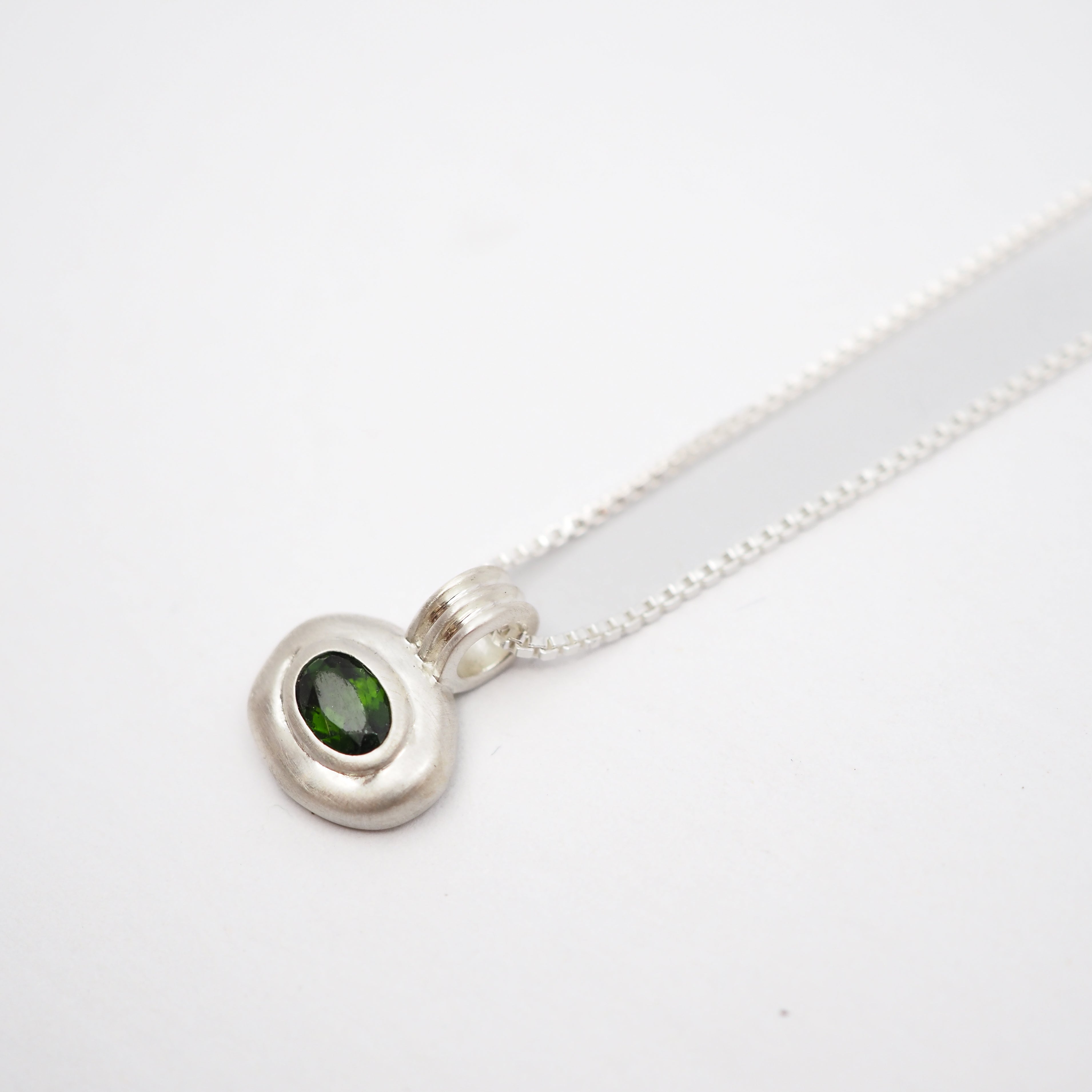 A sterling silver pendant necklace featuring a polished, organic-shaped silver bezel holding a round green gemstone, suspended from a delicate silver chain on a white background.