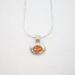 A silver pendant necklace featuring a textured silver bezel holding a small, oval-shaped orange gemstone, suspended from a delicate silver chain against a plain white background.