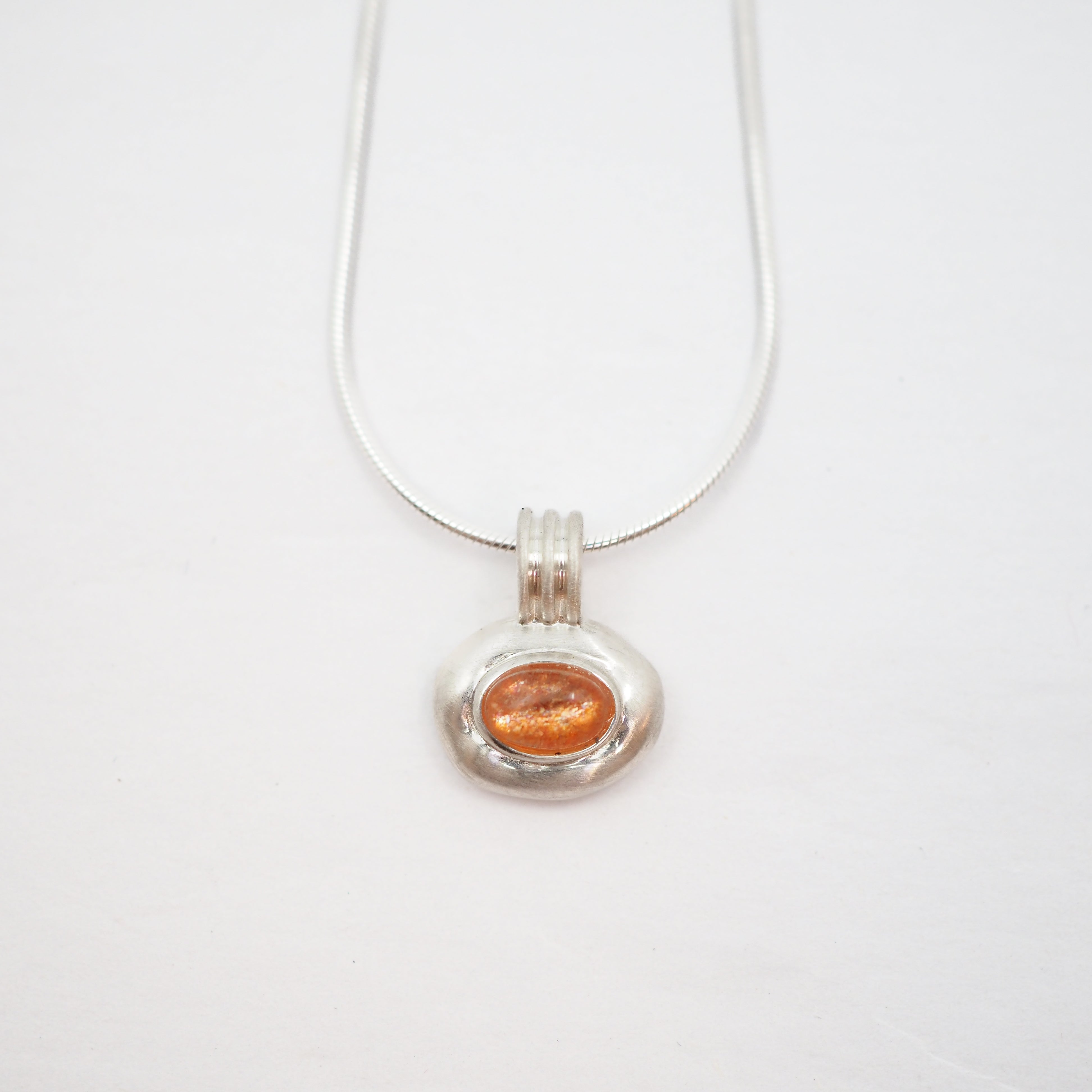 A silver pendant necklace featuring a textured silver bezel holding a small, oval-shaped orange gemstone, suspended from a delicate silver chain against a plain white background.