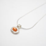 A silver pendant necklace featuring a textured, organic-shaped silver charm with a smooth, oval orange gemstone in the center, suspended from a thin silver snake chain on a white background.