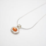 A silver pendant necklace featuring a textured, organic-shaped silver charm with a smooth, oval orange gemstone in the center, suspended from a thin silver snake chain on a white background.