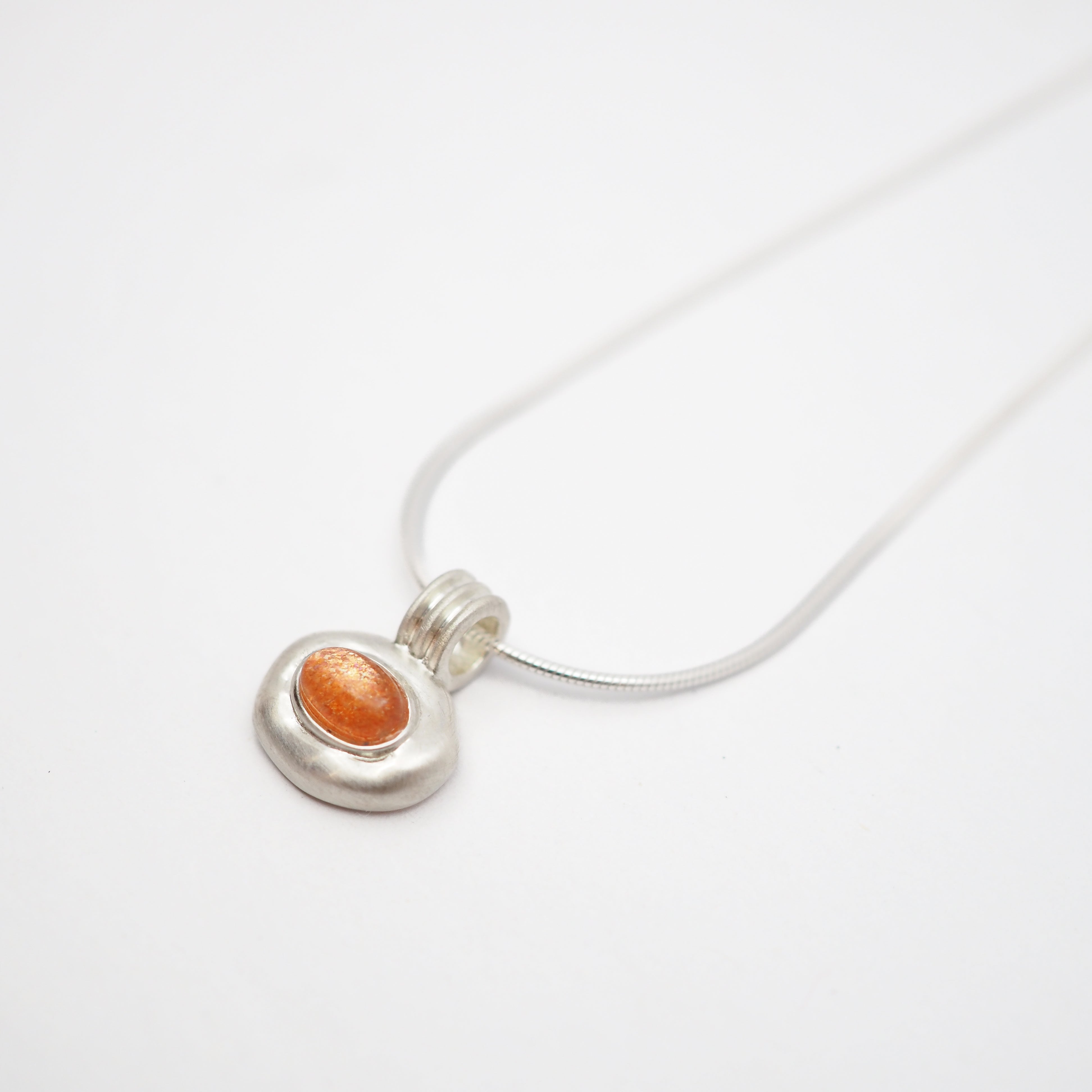 A silver pendant necklace featuring a textured, organic-shaped silver charm with a smooth, oval orange gemstone in the center, suspended from a thin silver snake chain on a white background.