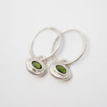 A pair of silver hoop earrings with small, irregular silver pendants set with oval green gemstones, shown on a plain white background.