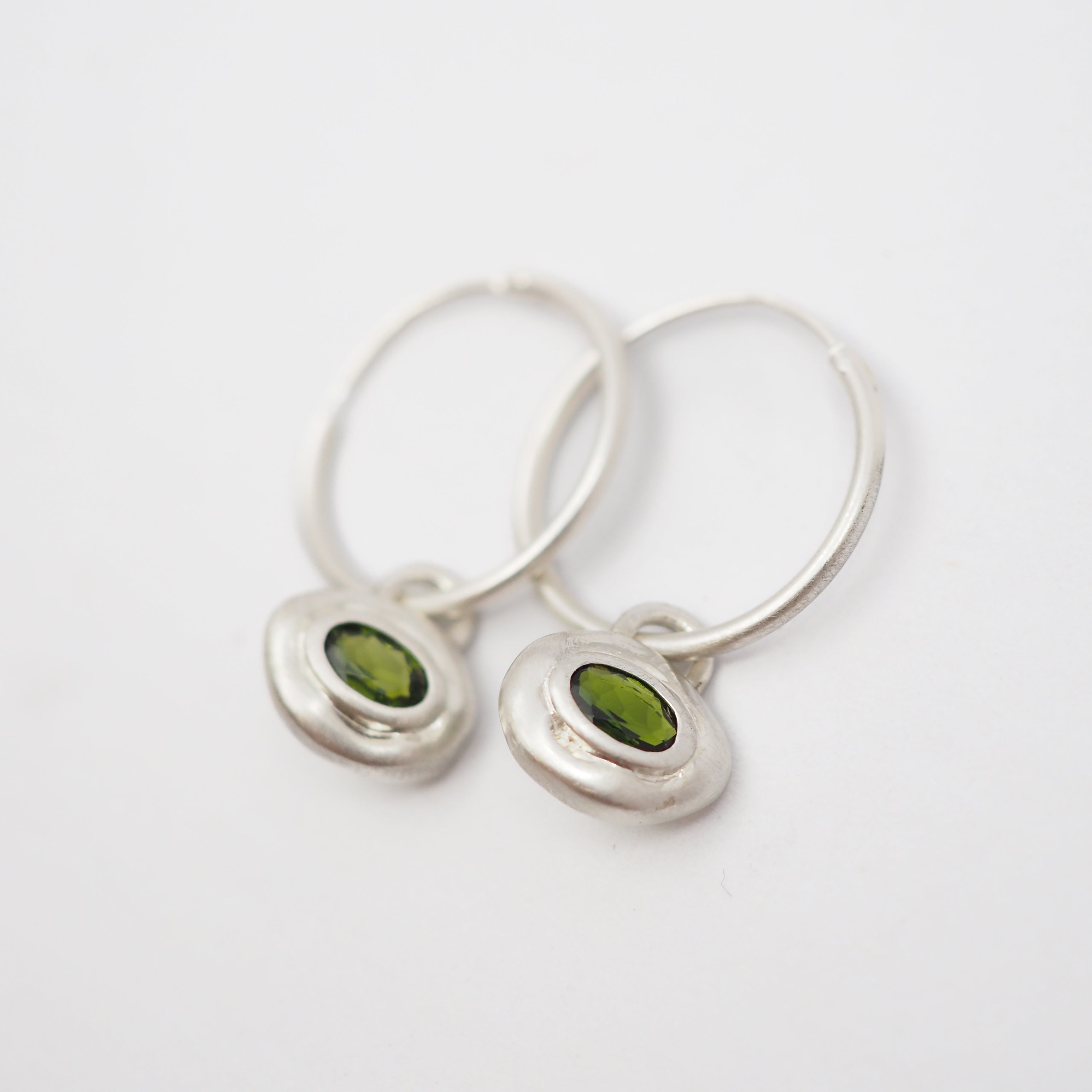 A pair of silver hoop earrings with small, irregular silver pendants set with oval green gemstones, shown on a plain white background.