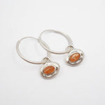 A pair of silver hoop earrings with small, bezel-set oval orange stones, displayed on a clean white background.