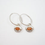 A pair of silver hoop earrings with small, irregular silver bezel settings holding round orange cabochon gemstones, displayed on a clean white background.