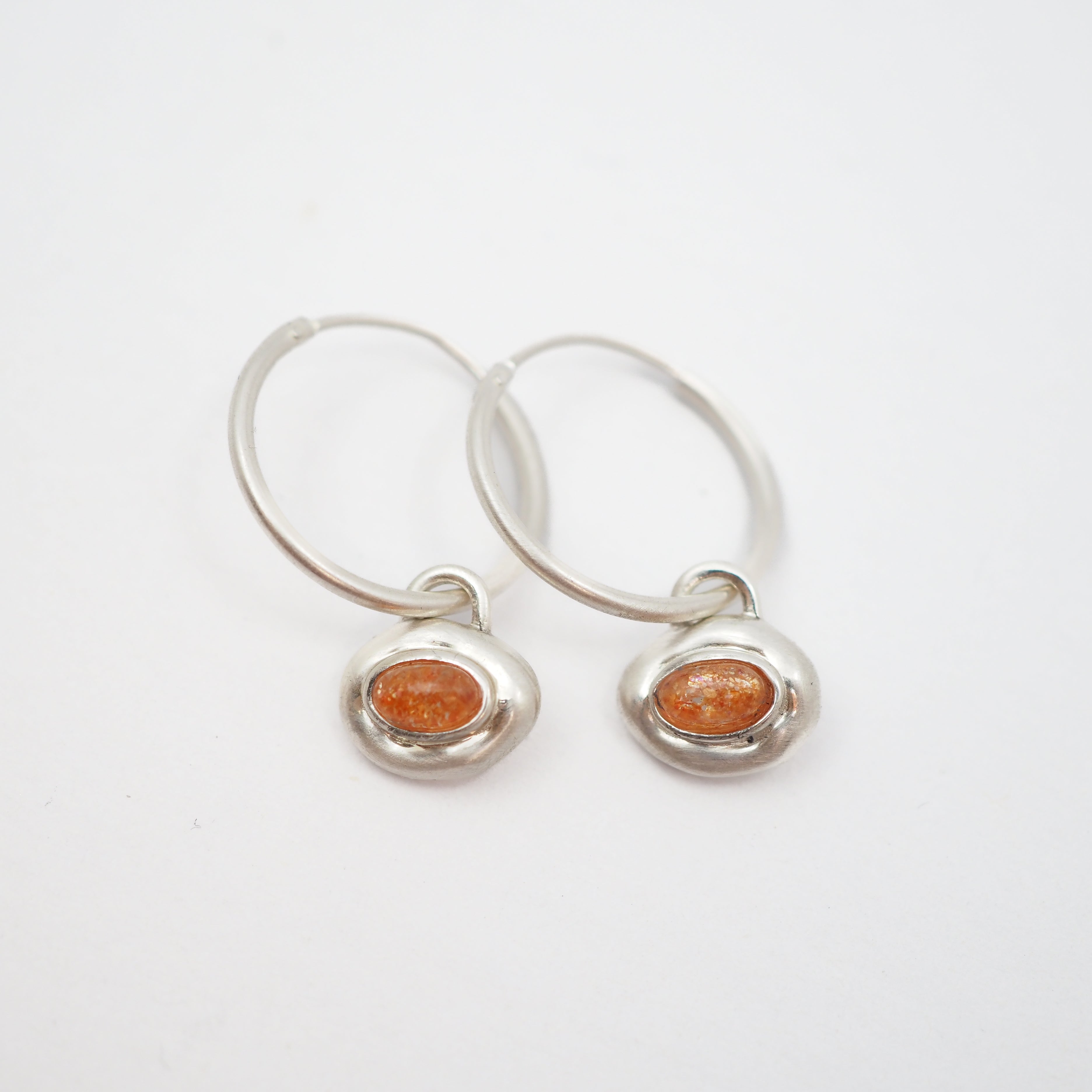 A pair of silver hoop earrings with small, irregular silver bezel settings holding round orange cabochon gemstones, displayed on a clean white background.