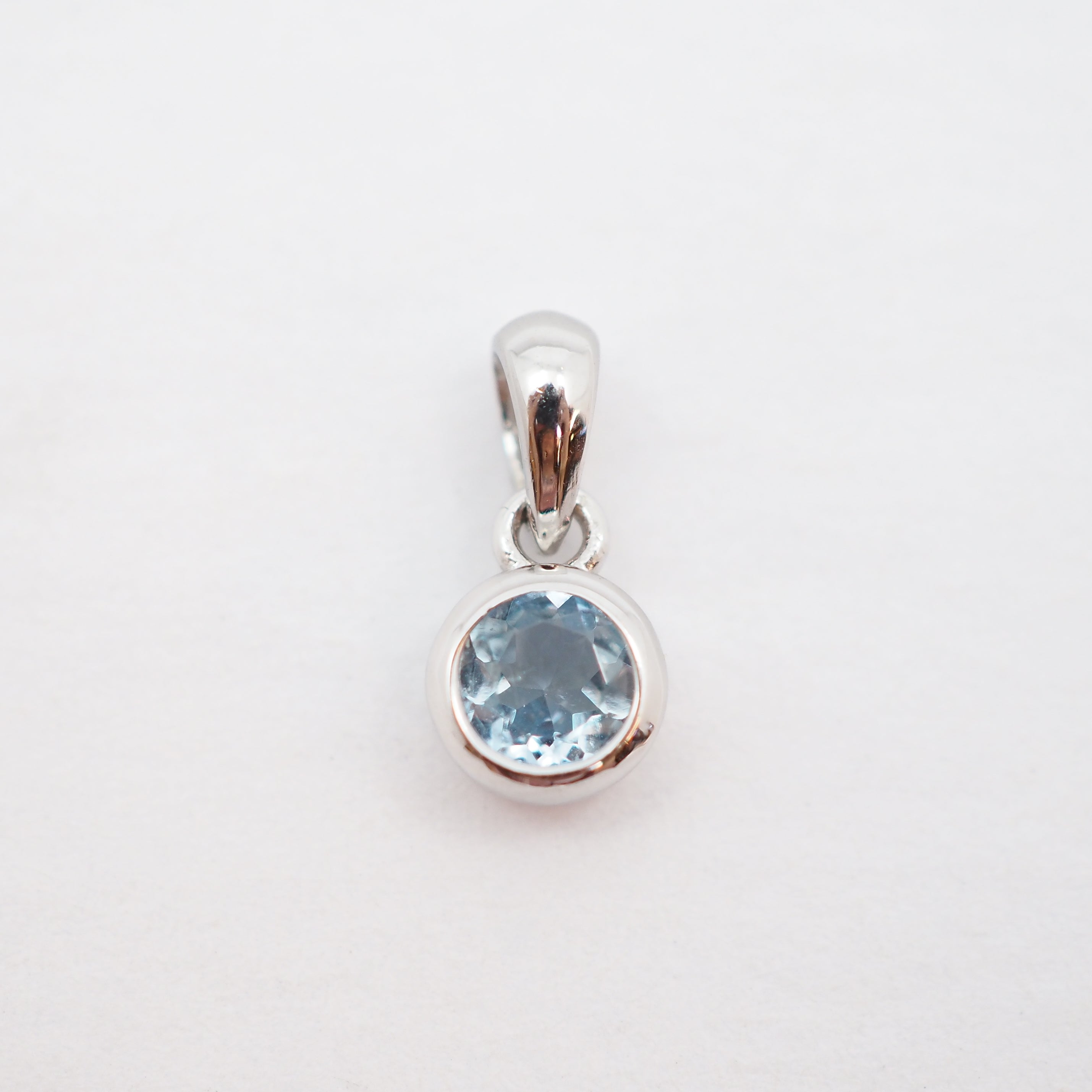A sterling silver pendant featuring a round-cut light blue gemstone in a bezel setting, suspended from a polished bail against a plain white background.