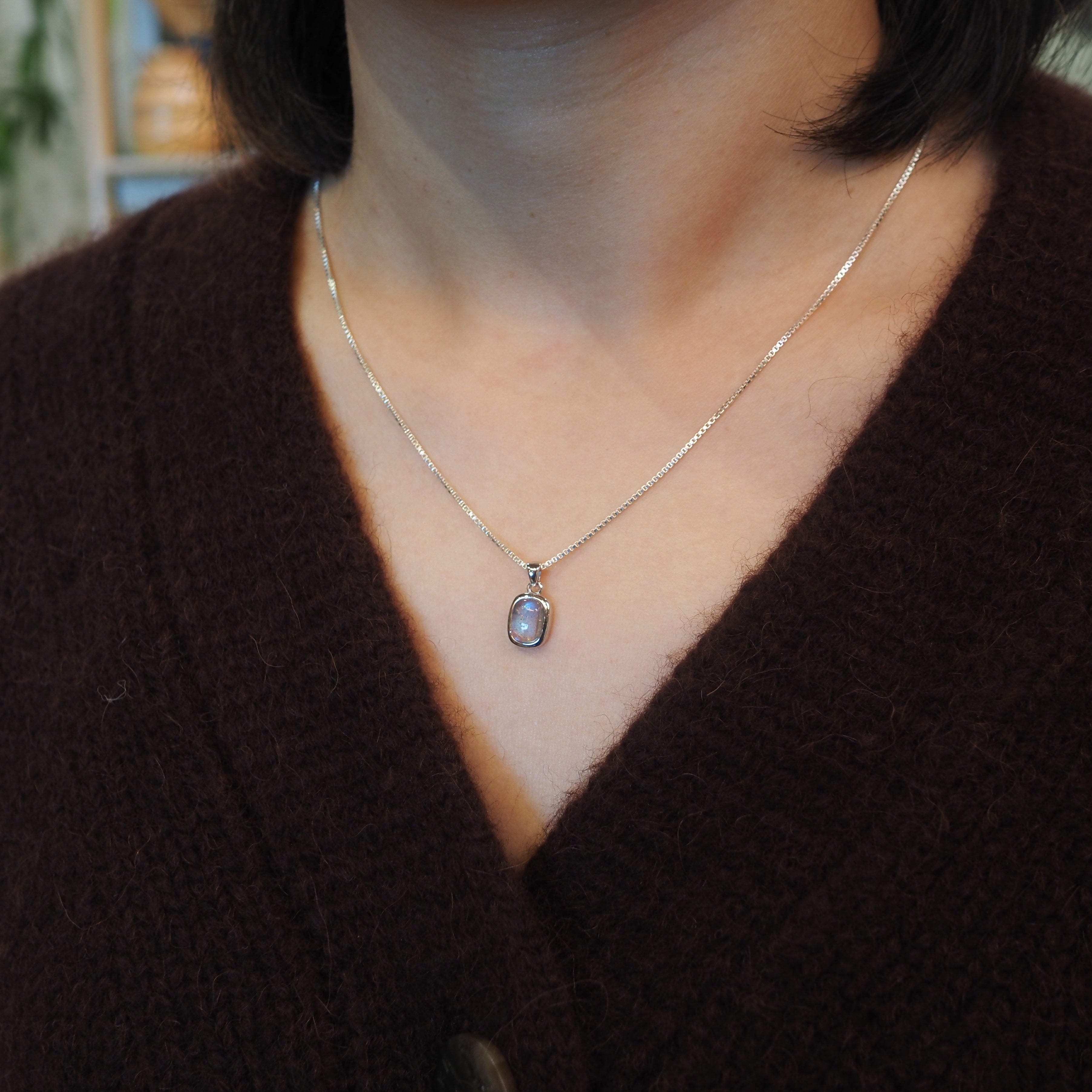 A close-up of a person wearing a delicate silver chain necklace with a small rectangular shaped pendant featuring a light blue-gray stone, styled with a dark brown knit cardigan.