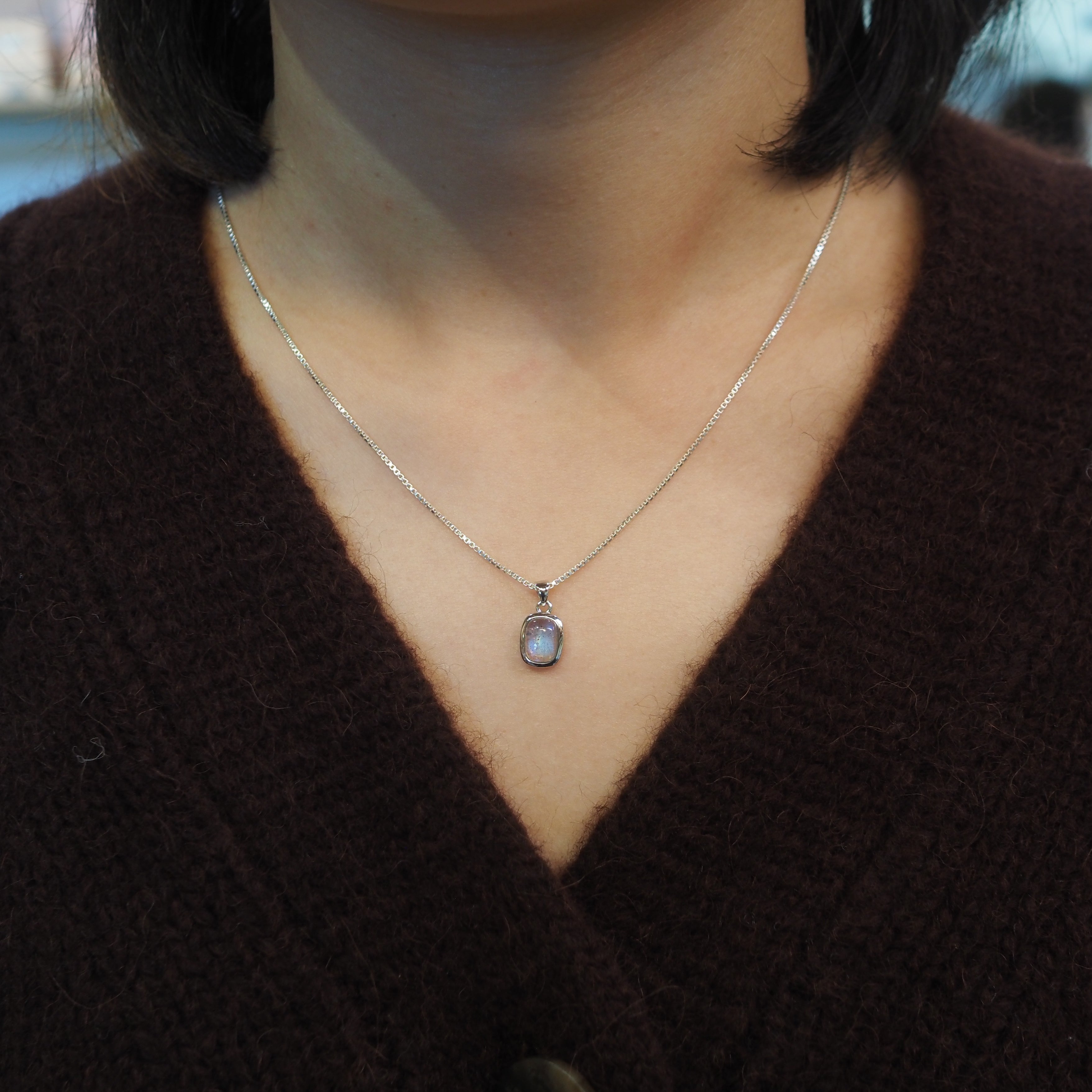 A close-up of a person wearing a delicate silver chain necklace with a small rectangular shaped pendant featuring a light blue-gray stone, styled with a dark brown knit cardigan.