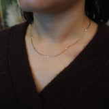 A close-up of a person wearing a delicate gold chain necklace with a textured, beaded appearance, styled with a dark brown V-neck sweater.