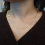 A close-up of a person's neck and chest wearing a delicate gold chain necklace over a dark brown knit sweater.