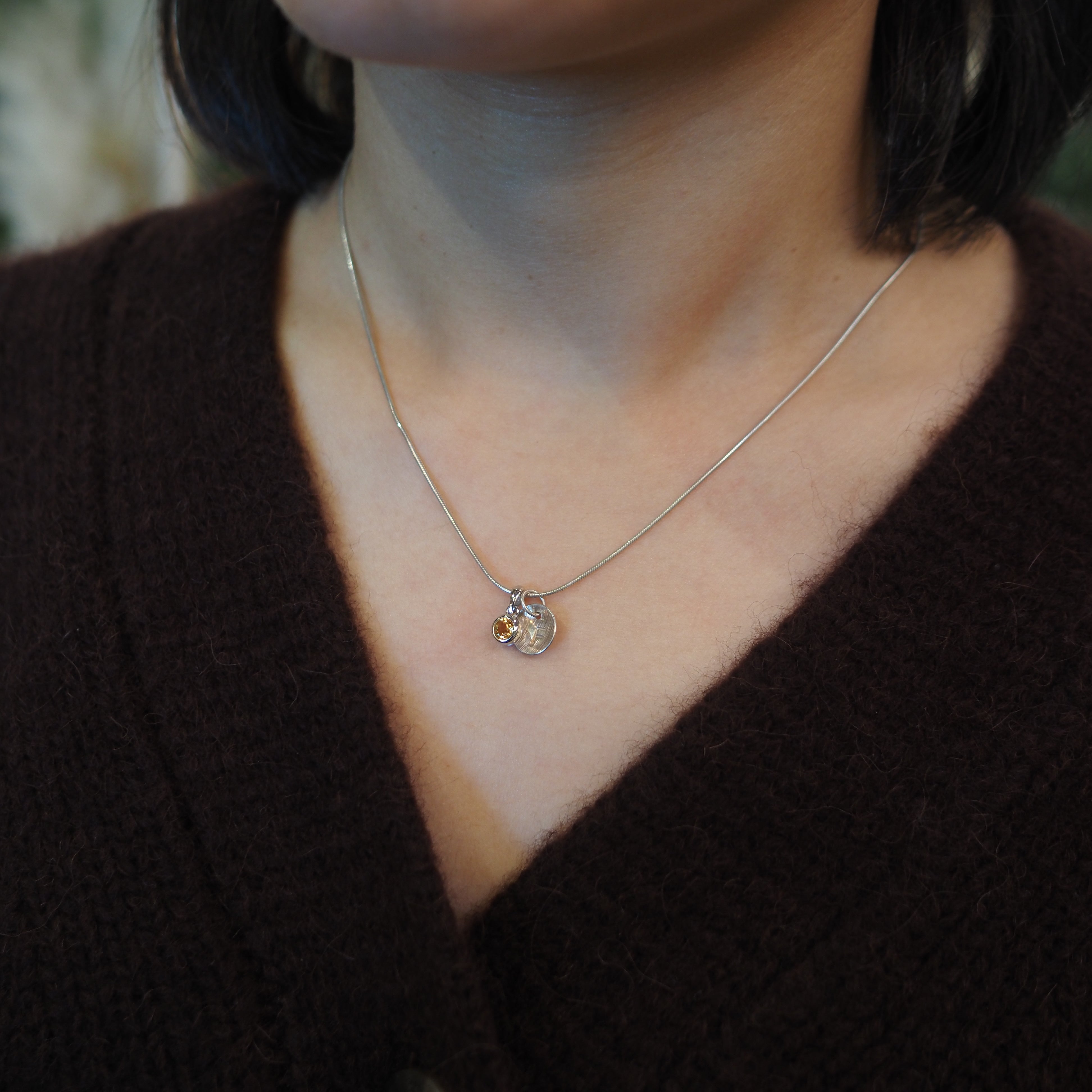 A close-up of a person wearing a delicate silver chain necklace with a small circular pendant featuring a tiny gold-toned accent, paired with a dark brown knit sweater.