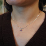 A close-up of a person wearing a delicate silver chain necklace with a small teardrop-shaped purple gemstone pendant, styled with a dark brown knit top.