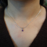 A close-up of a person's neck and chest wearing a delicate silver chain necklace with a small purple gemstone pendant.