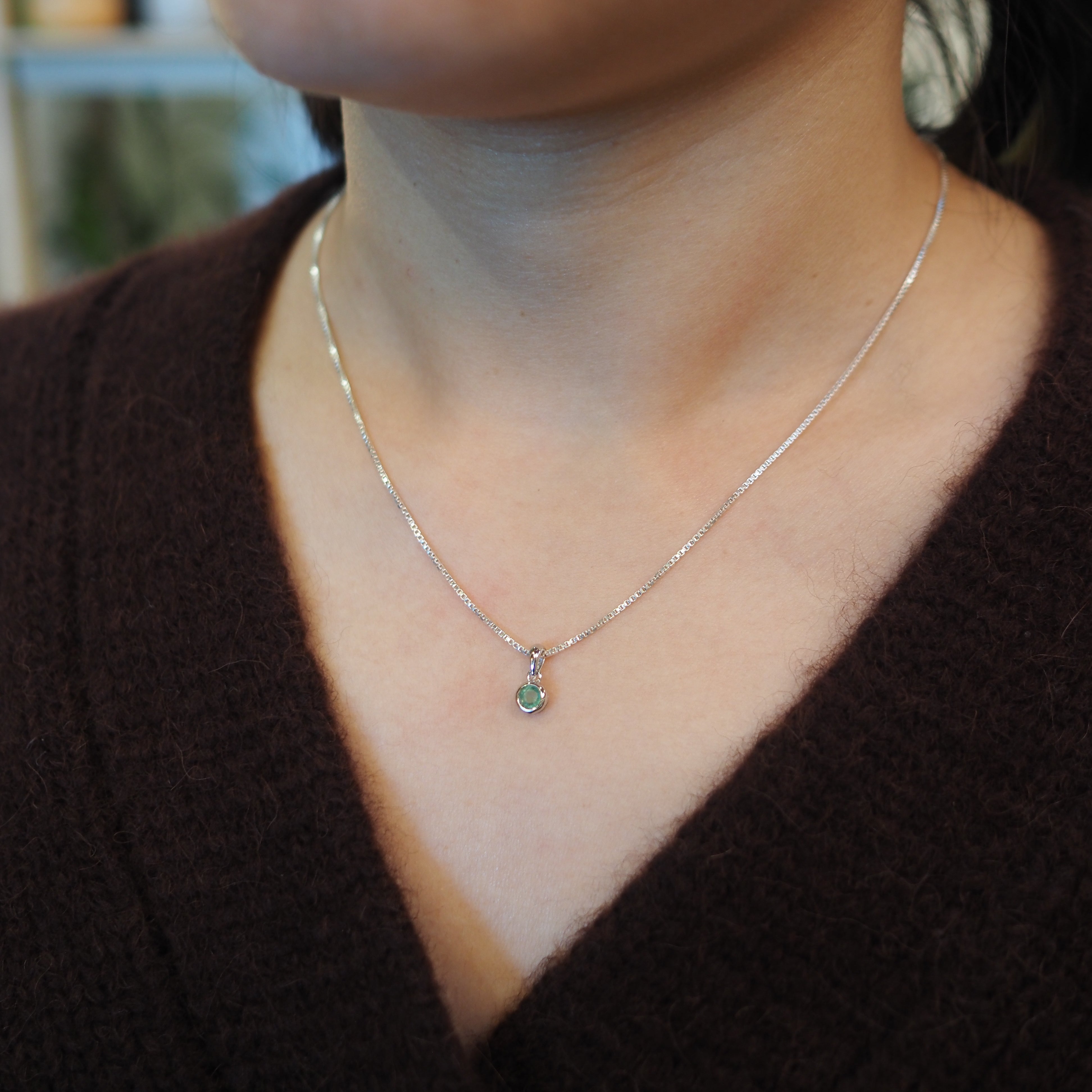 A close-up of a person wearing a delicate silver chain necklace with a small teardrop-shaped green gemstone pendant, styled with a dark brown knit sweater.