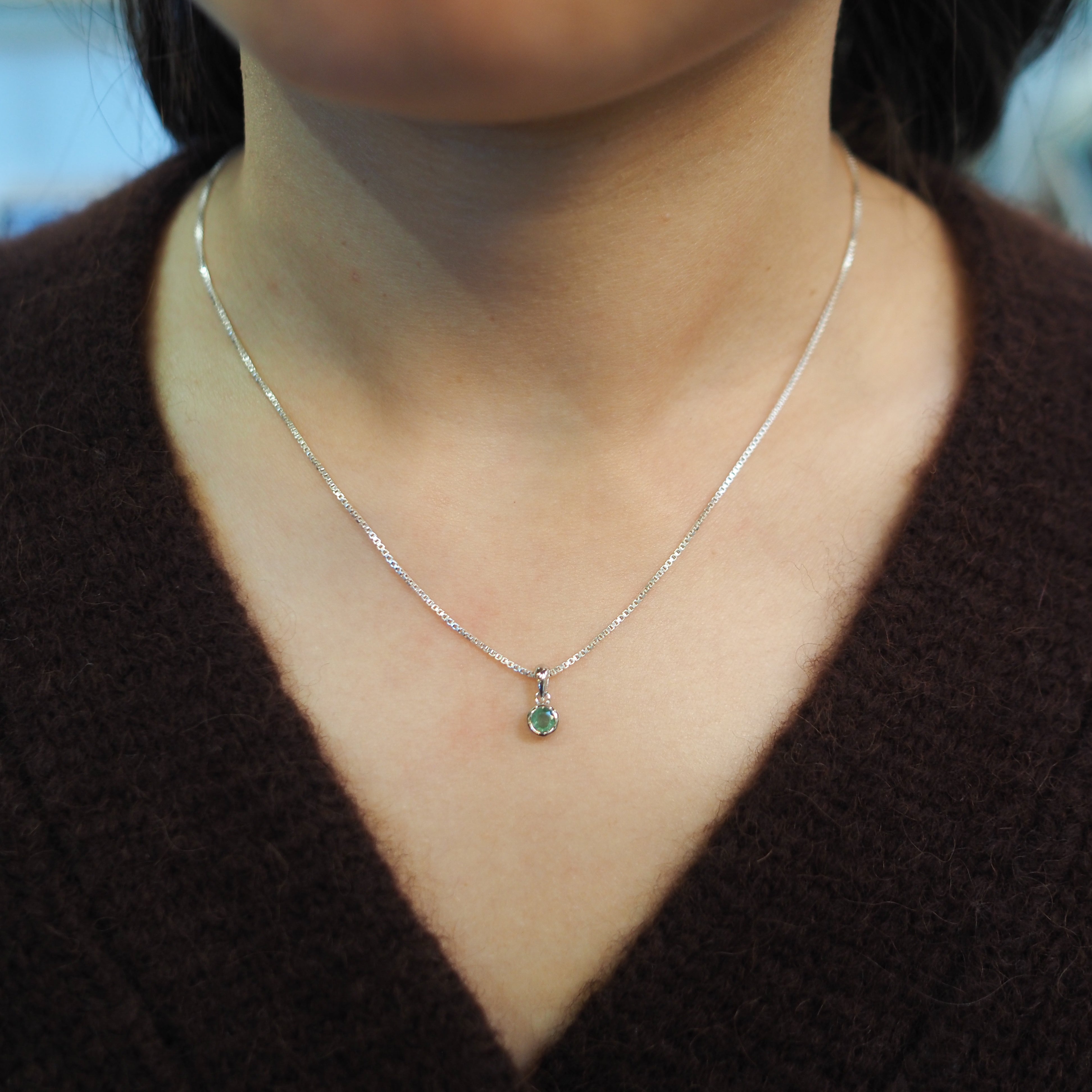 A close-up of a person's neck and chest wearing a delicate silver chain necklace with a small round green gemstone pendant, paired with a dark brown V-neck sweater.