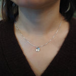 A close-up of a person wearing a delicate silver chain necklace with a small, polished silver disc pendant, styled with a dark brown knit top.