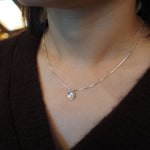 A close-up of a person's neck and chest wearing a delicate silver chain necklace with a small, faceted clear teardrop pendant.
