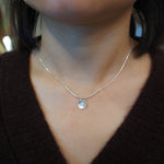 A close-up of a person's neck and chest wearing a delicate silver chain necklace with a small round silver pendant, paired with a dark brown knit top.
