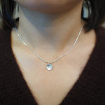 A close-up of a person's neck and chest wearing a delicate silver chain necklace with a small, round, silver pendant.