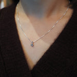 A close-up of a person wearing a delicate silver chain necklace with a small round clear gemstone pendant, styled with a dark brown knit sweater.