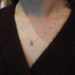 A close-up of a person wearing a delicate silver chain necklace with a small round clear gemstone pendant, styled with a dark brown knit sweater.