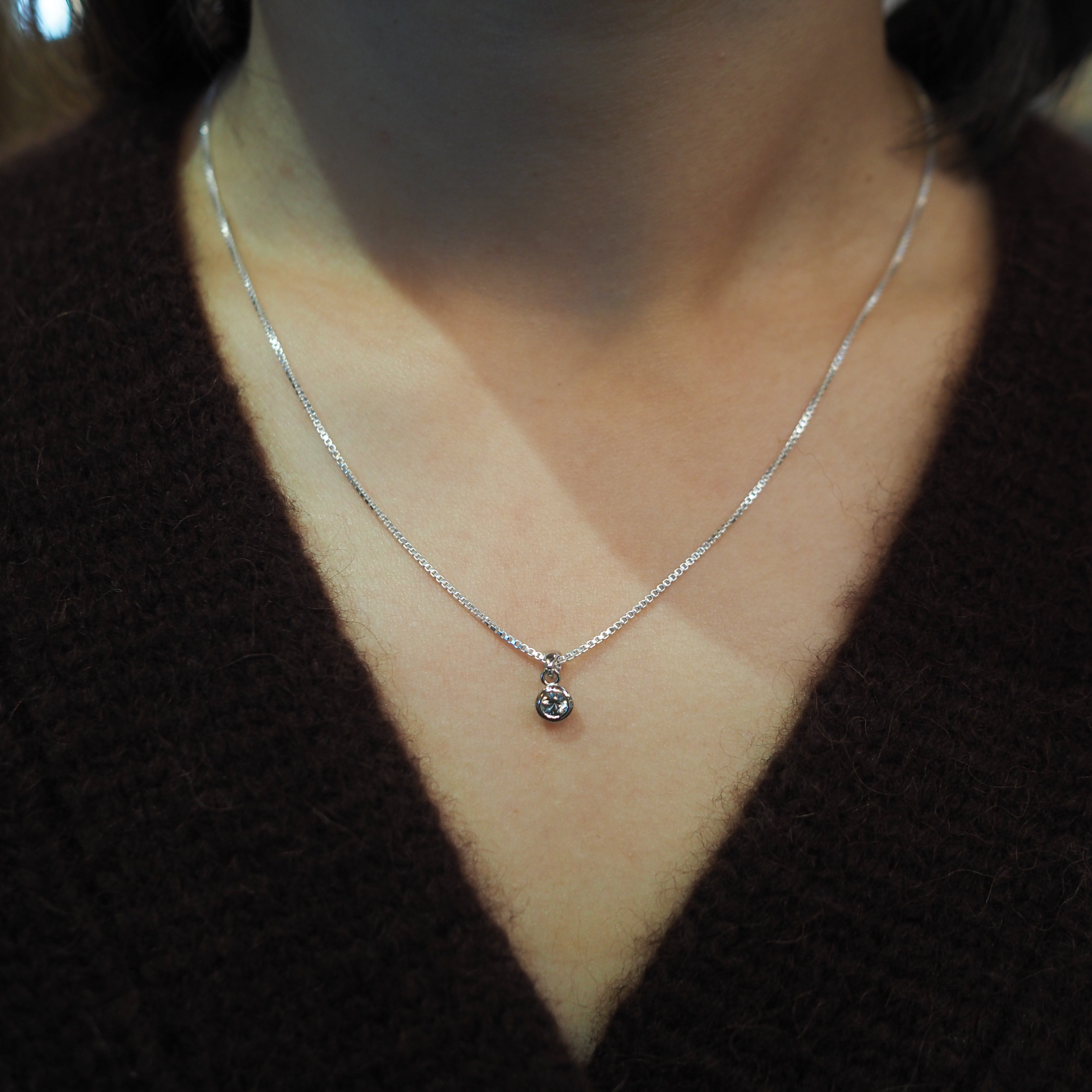 A close-up of a person wearing a delicate silver chain necklace with a small bezel-set clear gemstone pendant, styled over a dark brown V-neck sweater.