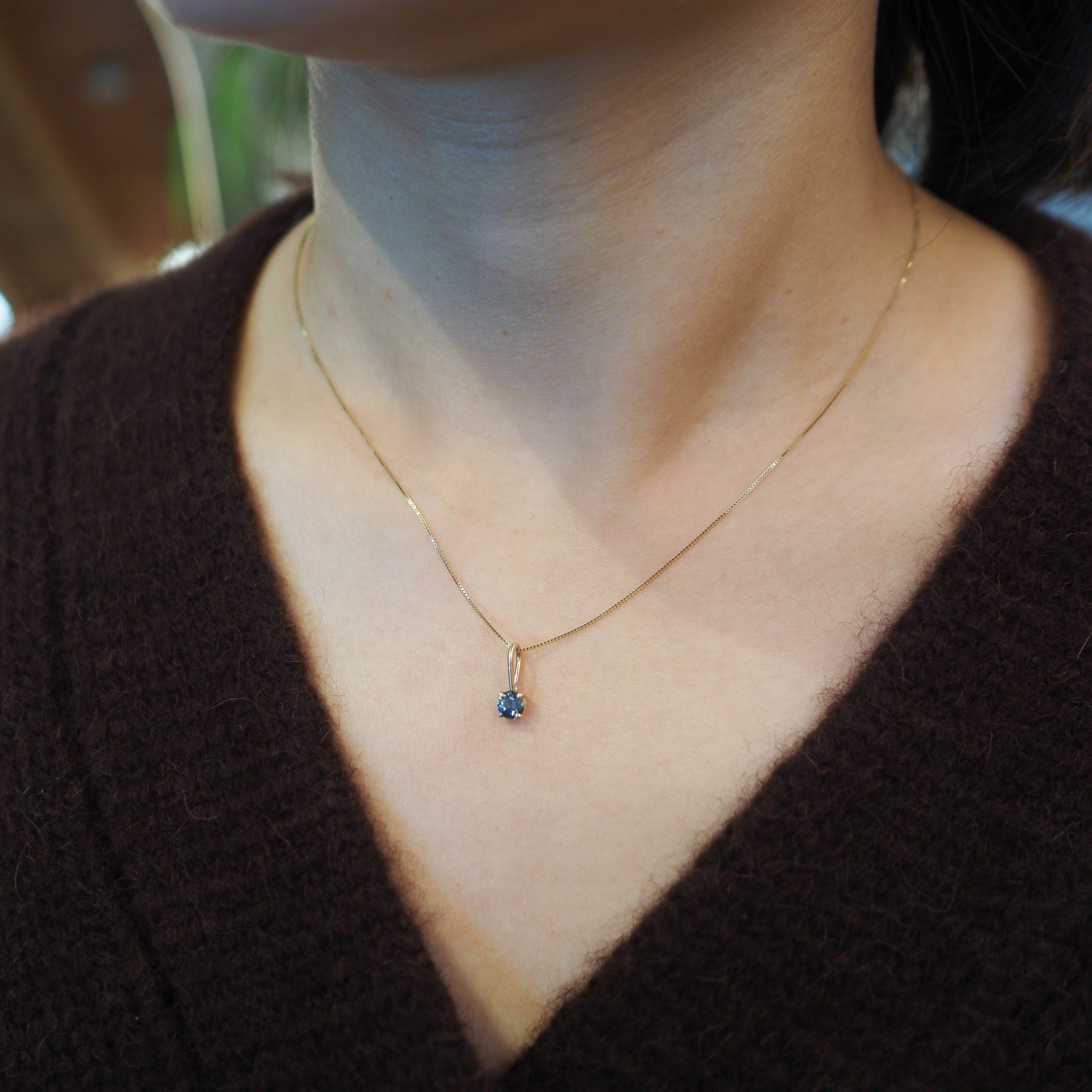 A close-up of a person's neck and chest wearing a delicate gold chain necklace with a small dark blue gemstone pendant, paired with a dark brown knit sweater.