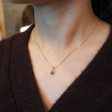 A close-up of a person's neck and chest wearing a delicate gold chain necklace with a small blue gemstone pendant, paired with a dark brown knit top.