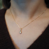A delicate gold chain necklace with a small oval pendant worn on a person's neck over a dark brown knit top.