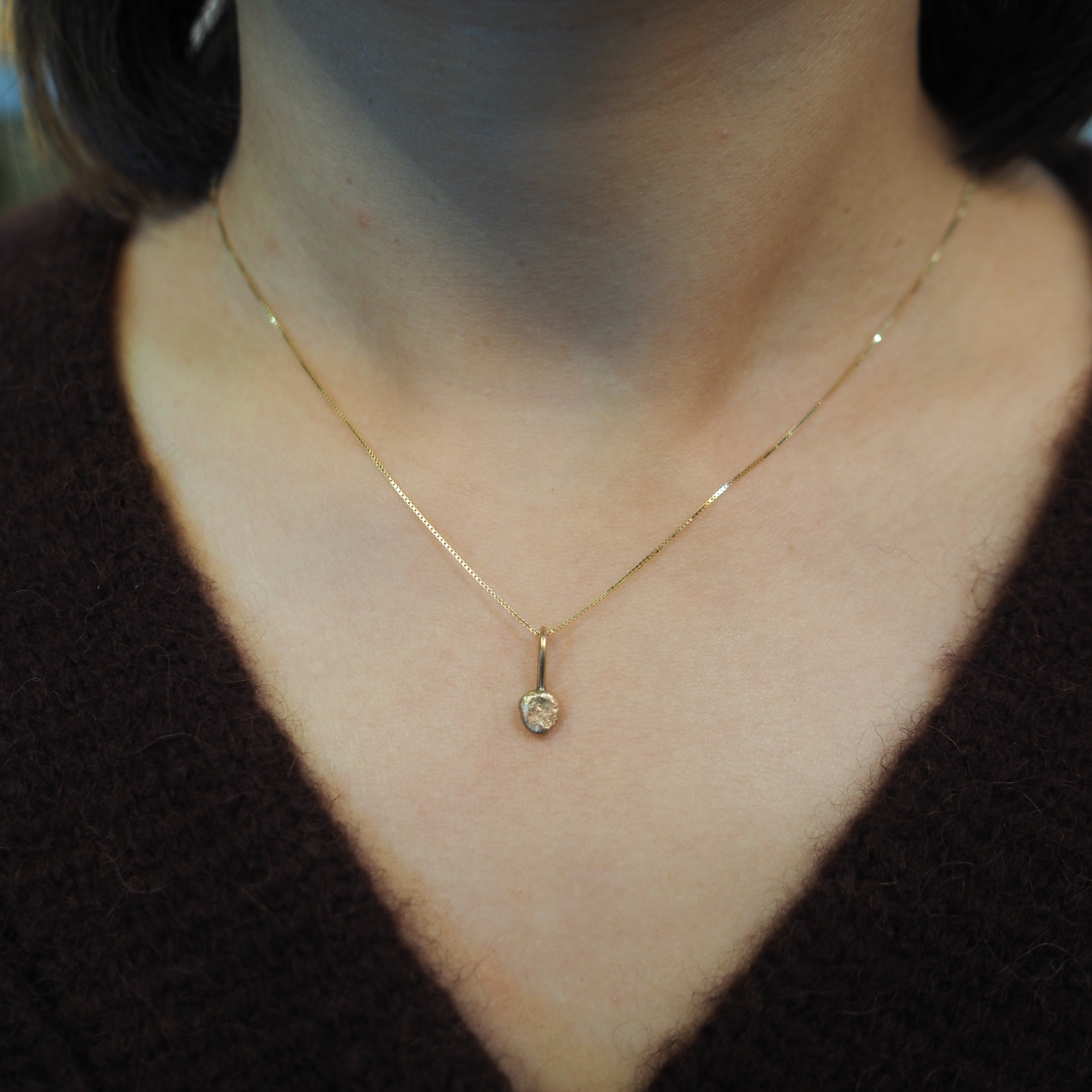 A delicate gold chain necklace with a small round pendant, shown worn on a person's neck over a dark brown knit top.