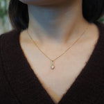 A close-up of a person's neck and chest wearing a delicate gold chain necklace with a small pendant, paired with a dark brown knit V-neck sweater.