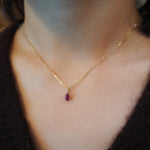 A delicate gold chain necklace with a small oval red gemstone pendant, worn on a person's neck over a dark brown knit top.