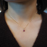 A close-up of a person's neck and chest wearing a delicate gold chain necklace with a small oval-shaped dark red gemstone pendant.