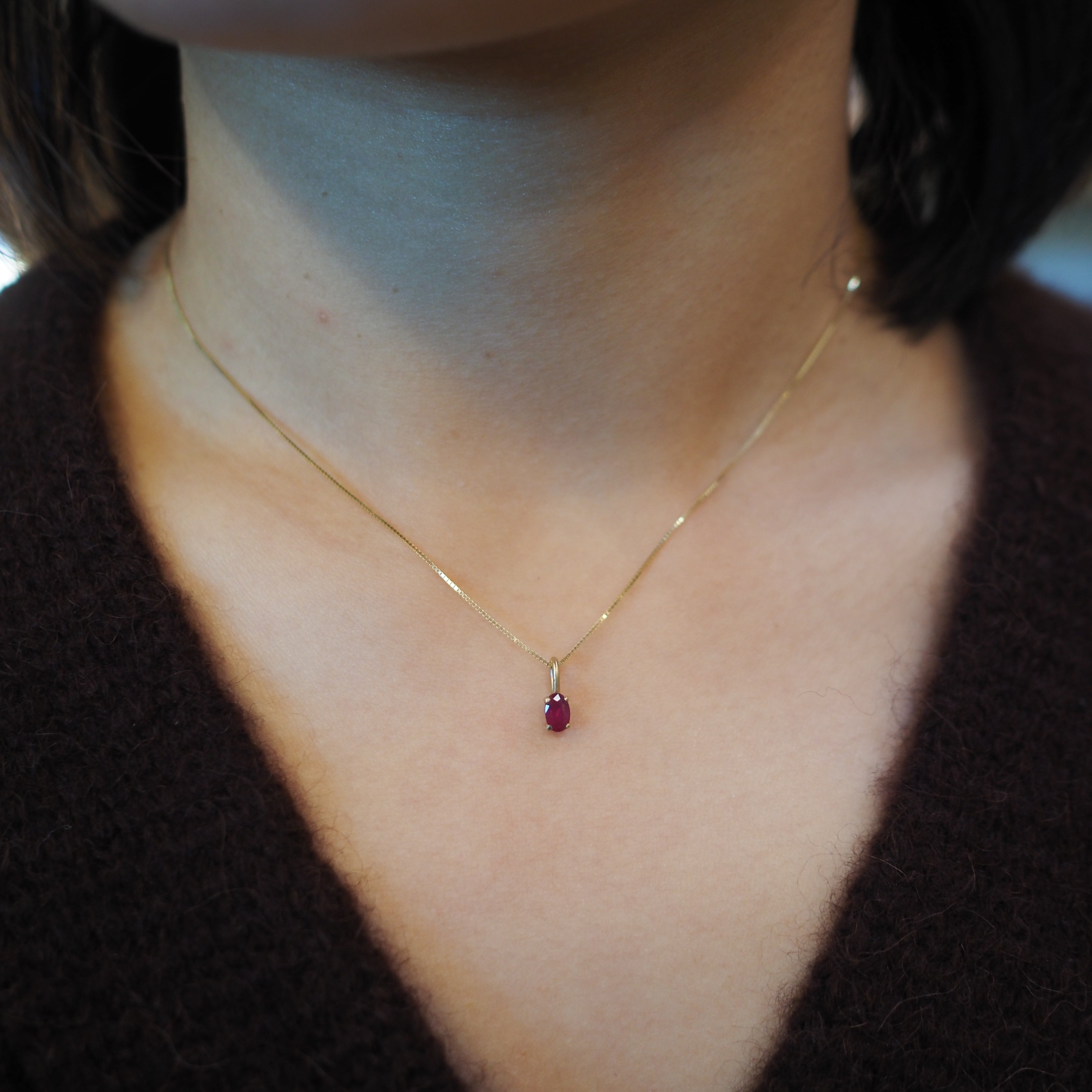 A close-up of a person's neck and chest wearing a delicate gold chain necklace with a small oval-shaped dark red gemstone pendant.