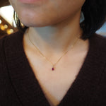 A close-up of a person wearing a delicate gold chain necklace with a small oval-shaped dark red gemstone pendant.
