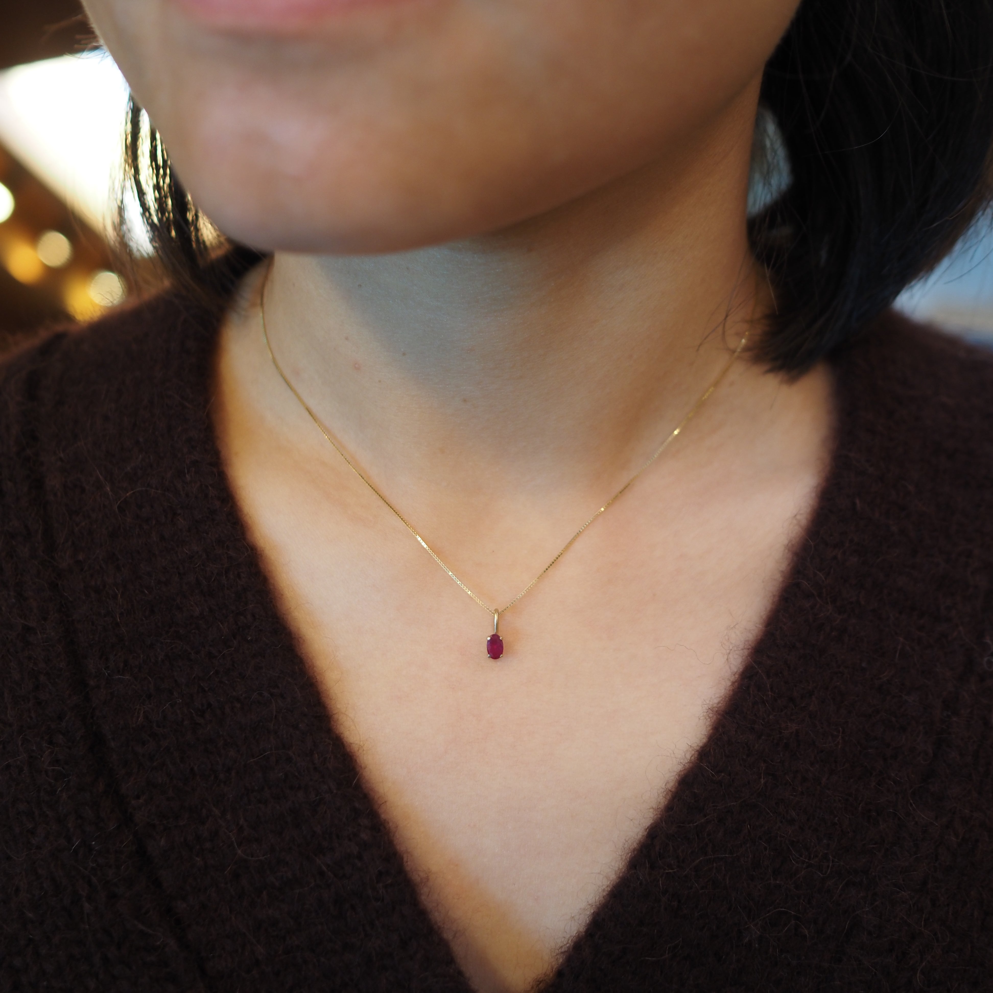 A close-up of a person wearing a delicate gold chain necklace with a small oval-shaped dark red gemstone pendant.
