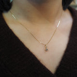A close-up of a person's neck and chest wearing a delicate gold chain necklace with a small teardrop-shaped gold pendant featuring a clear gemstone, paired with a dark brown knit top.