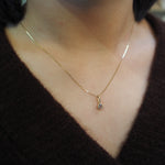 A close-up of a person's neck and chest wearing a delicate gold chain necklace with a small teardrop-shaped gold pendant featuring a clear gemstone, paired with a dark brown knit top.