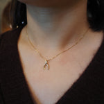 A close-up of a person's neck and chest wearing a delicate gold chain necklace with a small gold wishbone pendant, paired with a dark brown knit sweater.