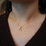 A close-up of a person's neck and chest wearing a delicate gold chain necklace with a small gold wishbone pendant, paired with a dark brown knit sweater.