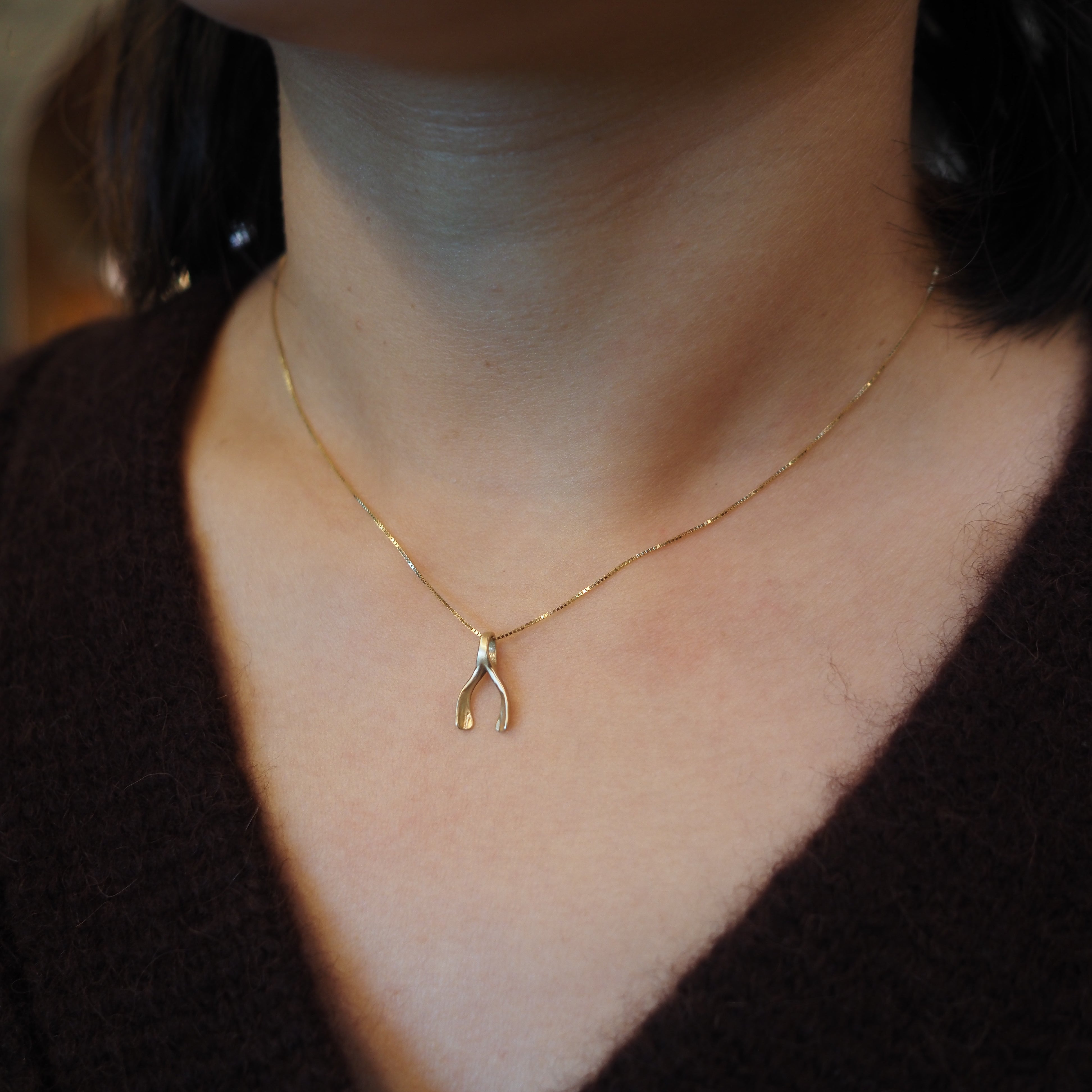 A close-up of a person's neck and chest wearing a delicate gold chain necklace with a small gold wishbone pendant, paired with a dark brown knit sweater.