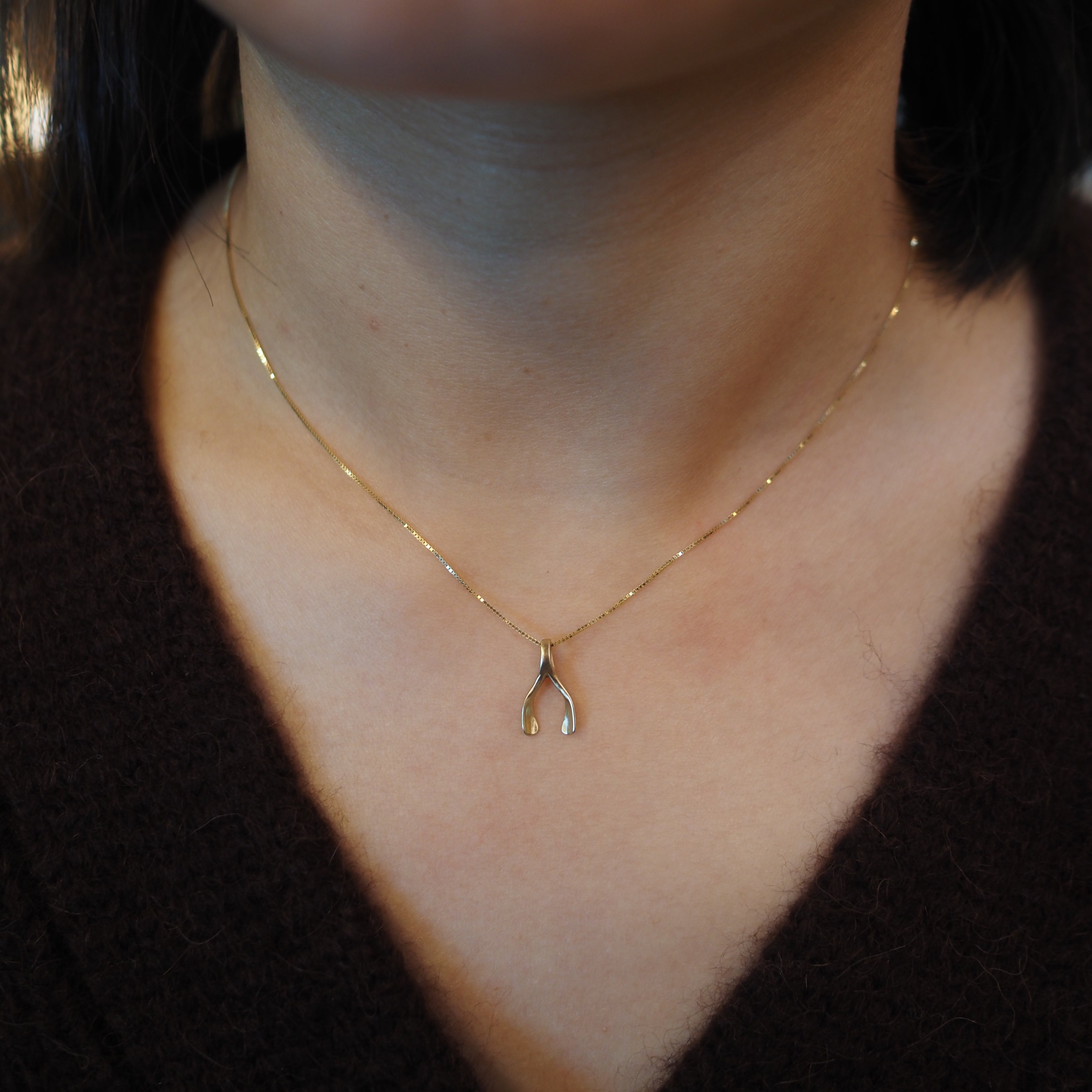 A close-up of a person's neck and chest wearing a delicate gold chain necklace with a small wishbone pendant.