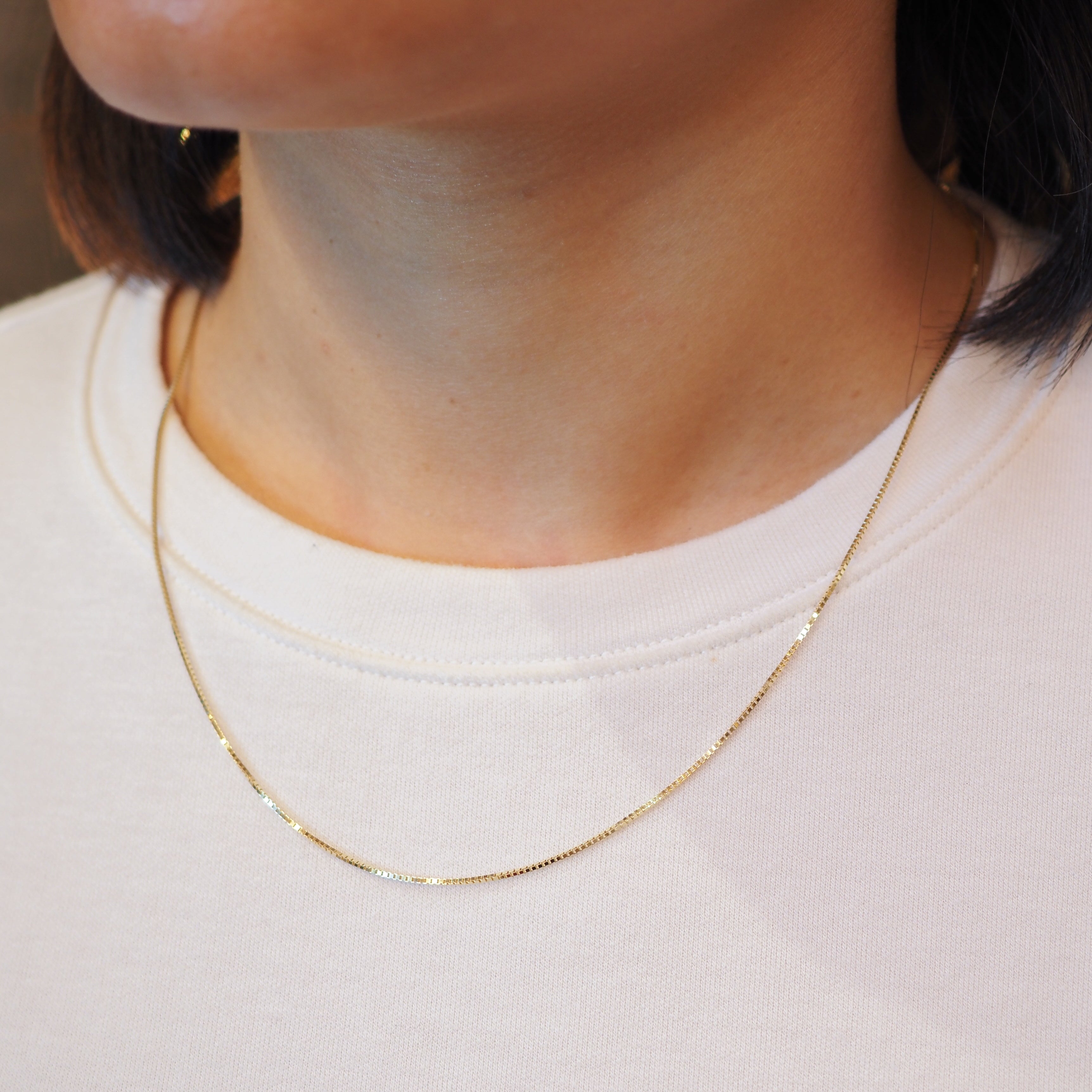 A close-up of a person's neck and upper chest wearing a delicate gold chain necklace over a white crewneck top.