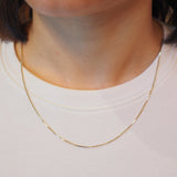 A close-up of a person's neck and upper chest wearing a delicate gold chain necklace over a white crew-neck t-shirt.